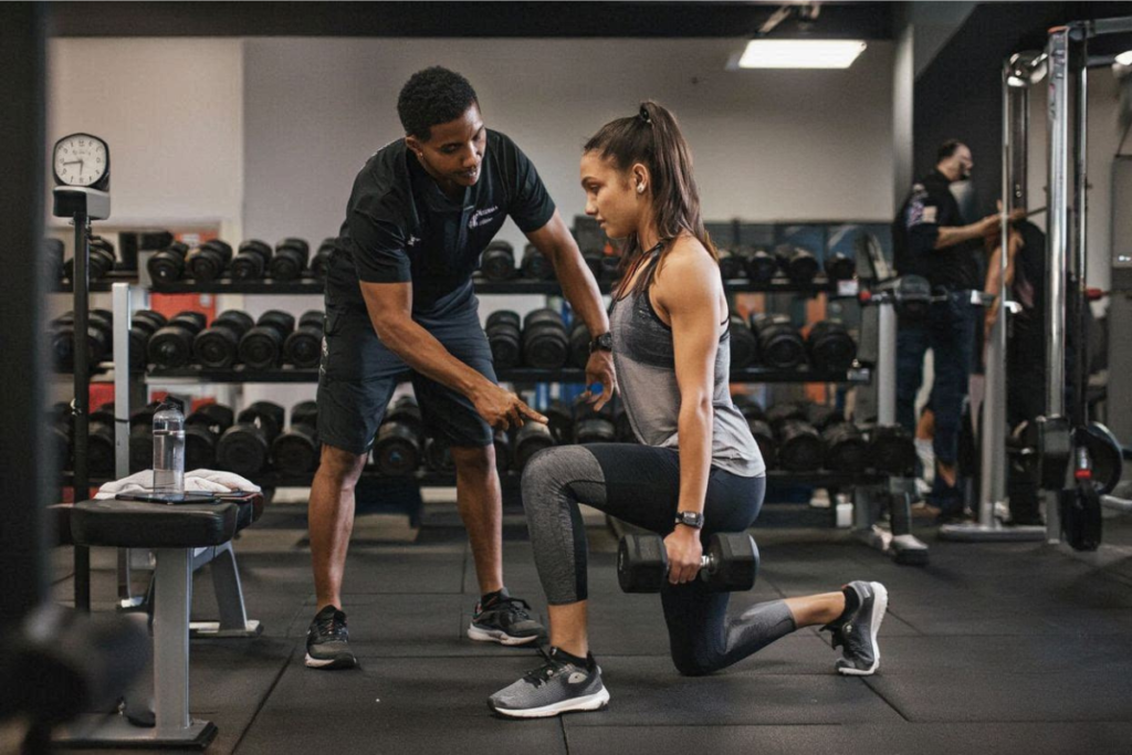 Una joven haciendo ejercicio en el gimnasio con mancuernas rodilla derecha flexionada contra el suelo recibiendo consejos de otro joven